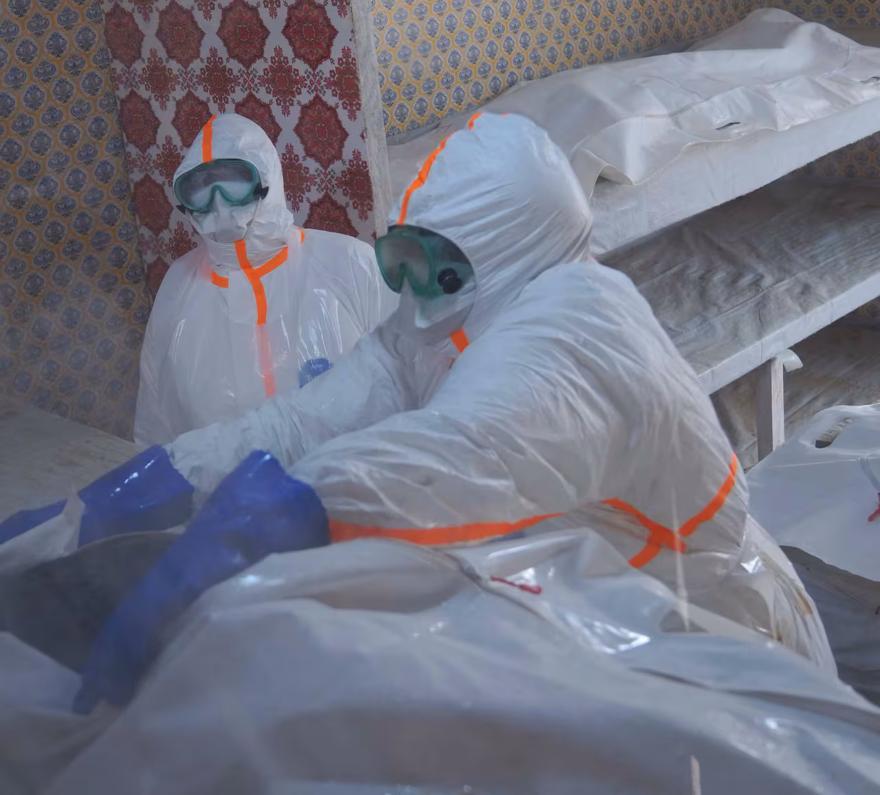 Two people in full protective gear in a room with people in body bags on beds.