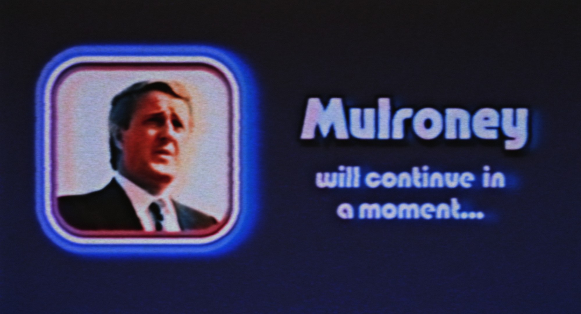A pixelated screen features a square with a man in a suit shot from the chest up. Next to it in blue text reads “Mulroney will continue in a moment” against a black background.
