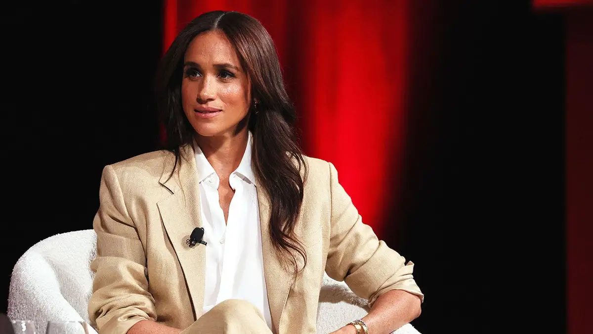 Meghan Markle speaks on stage in a tan suit and white shirt.